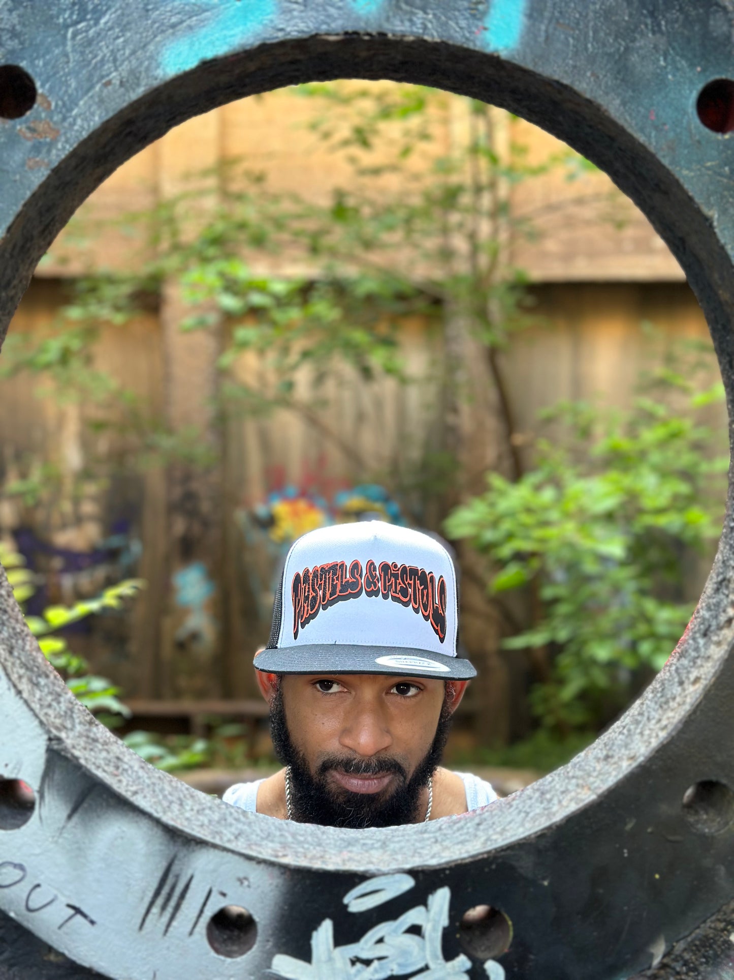 
@jaethejewelerugz in the Pastels & Pistols Mother Puffer Flat Bill Trucker Hat. White-and-red flat bill trucker with embroidered lettering. Lifestyle outdoor shot with natural greenery.