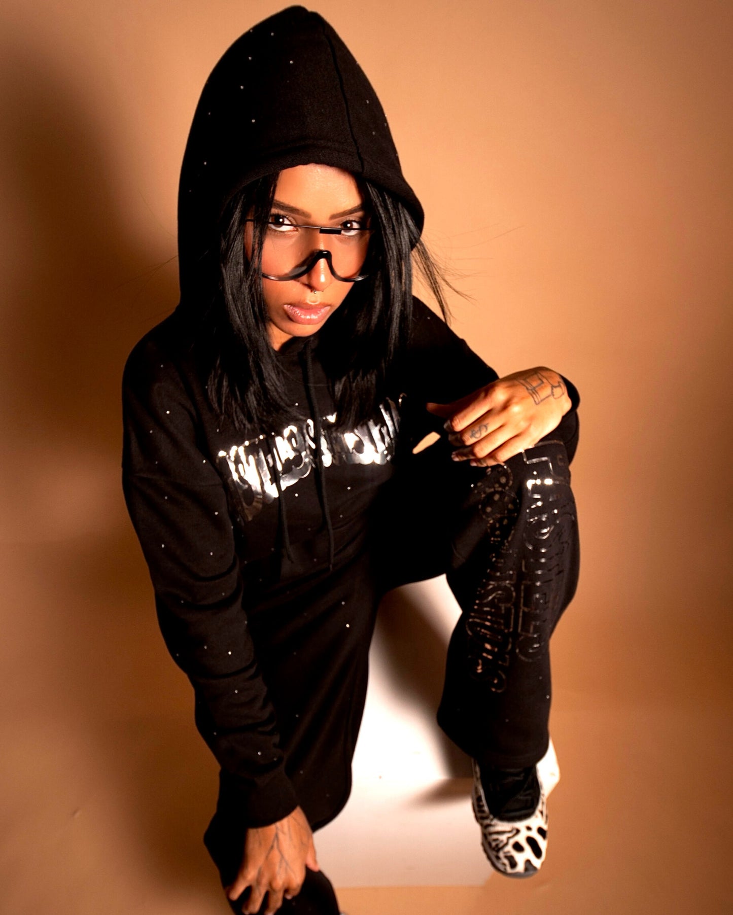 @yoselinpacheco394 wearing the Pastels & Pistols MotionDoLL Black Rhinestone Sweatsuit. Full-body angle showing hoodie and joggers with chrome vinyl text and rhinestones. Warm studio shot. Womens luxury sweatsuit.