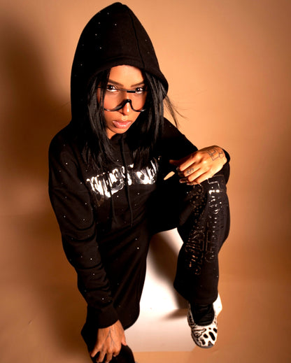 @yoselinpacheco394 wearing the Pastels & Pistols MotionDoLL Black Rhinestone Sweatsuit. Full-body angle showing hoodie and joggers with chrome vinyl text and rhinestones. Warm studio shot. Womens luxury sweatsuit.