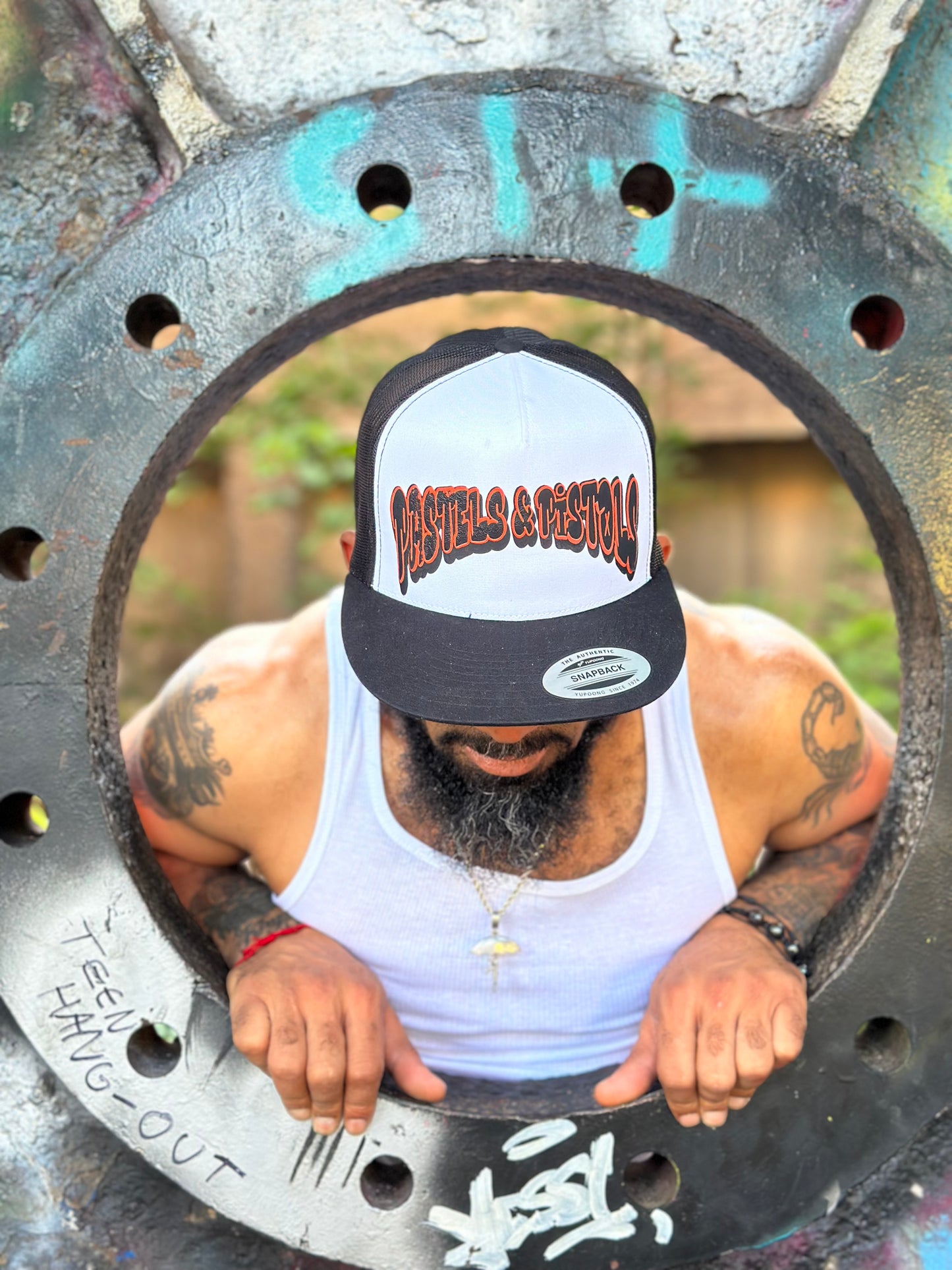 Close-up of @jaethejewelerugz wearing the Pastels & Pistols Mother Puffer Flat Bill Trucker Hat. White mesh-back trucker with red embroidery. Industrial outdoor background.