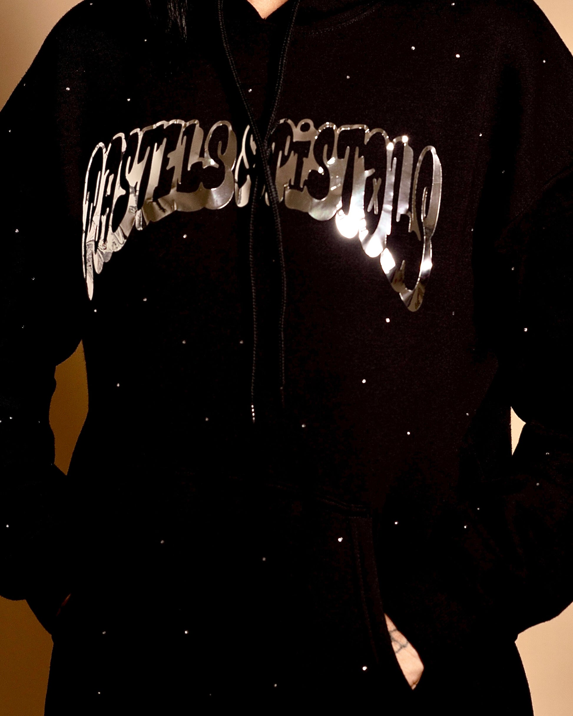 Close-up of the Pastels & Pistols MotionDoLL Black Rhinestone Hoodie. Chrome vinyl logo and rhinestone detailing on the front chest. Warm studio backdrop. Womens luxury streetwear.