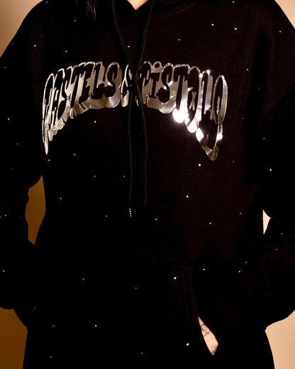 Close-up of the Pastels & Pistols MotionDoLL Black Rhinestone Hoodie. Chrome vinyl logo and rhinestone detailing on the front chest. Warm studio backdrop. Womens luxury streetwear.