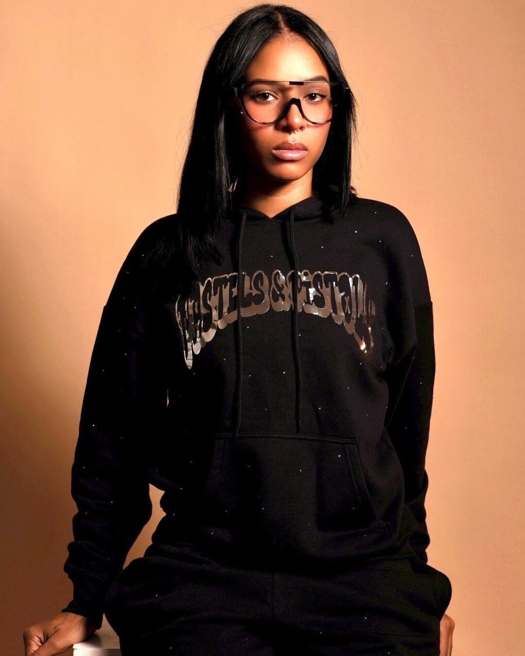 @yoselinpacheco394 in the Pastels & Pistols MotionDoLL Black Rhinestone Hoodie. Straight-on chest view highlighting chrome vinyl and rhinestones. Warm studio backdrop. Womens streetwear hoodie.
