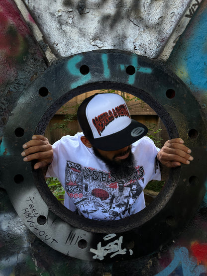 @jaethejewelerugz in the Pastels & Pistols Mother Puffer Flat Bill Trucker Hat. White-and-red trucker with adjustable snapback. Shot inside a large metal pipe for urban streetwear styling.
