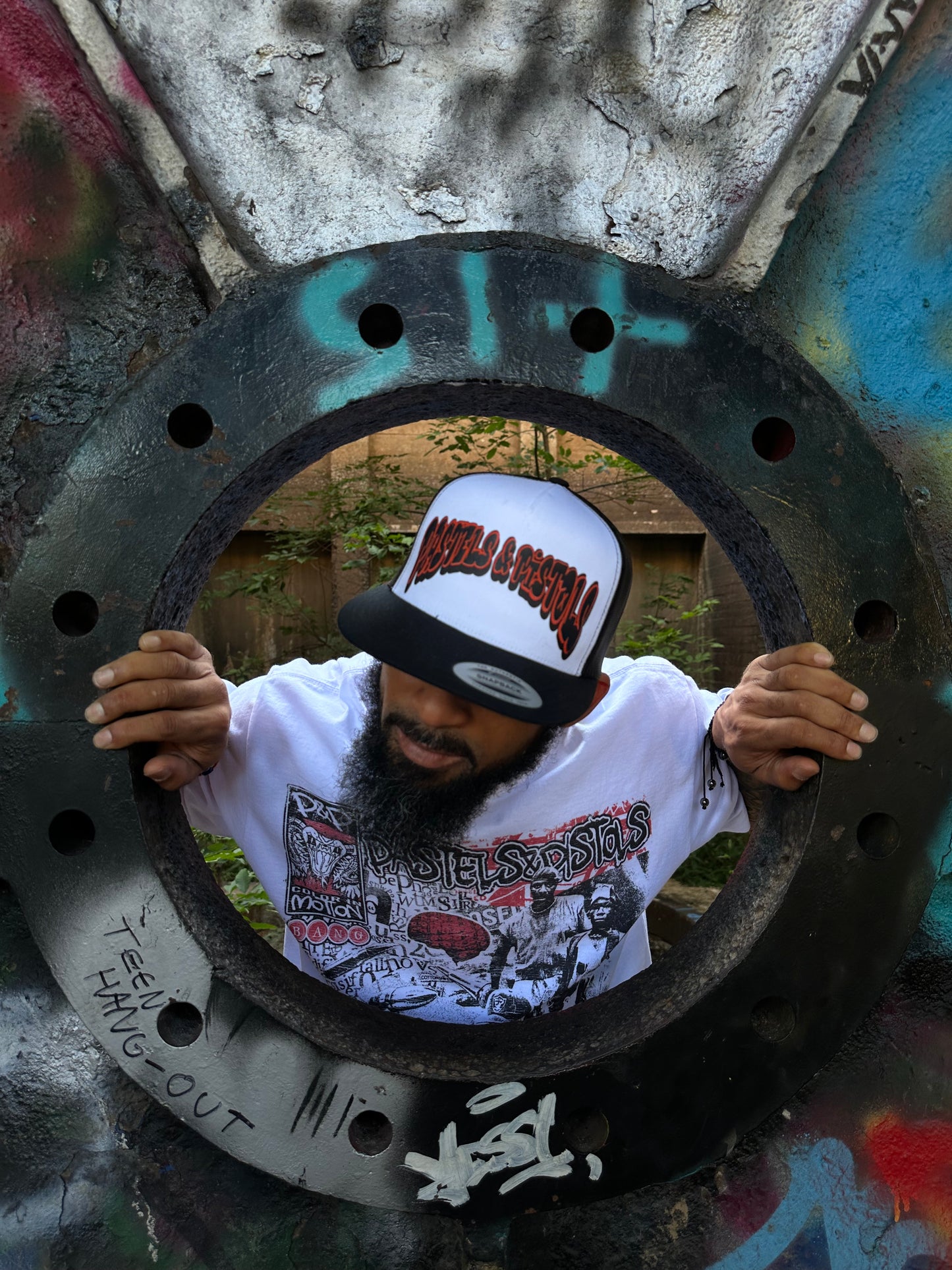 @jaethejewelerugz wearing the Pastels & Pistols Mother Puffer Flat Bill Trucker Hat. White front panel with red embroidered lettering. Outdoor industrial pipe setting for streetwear product display.