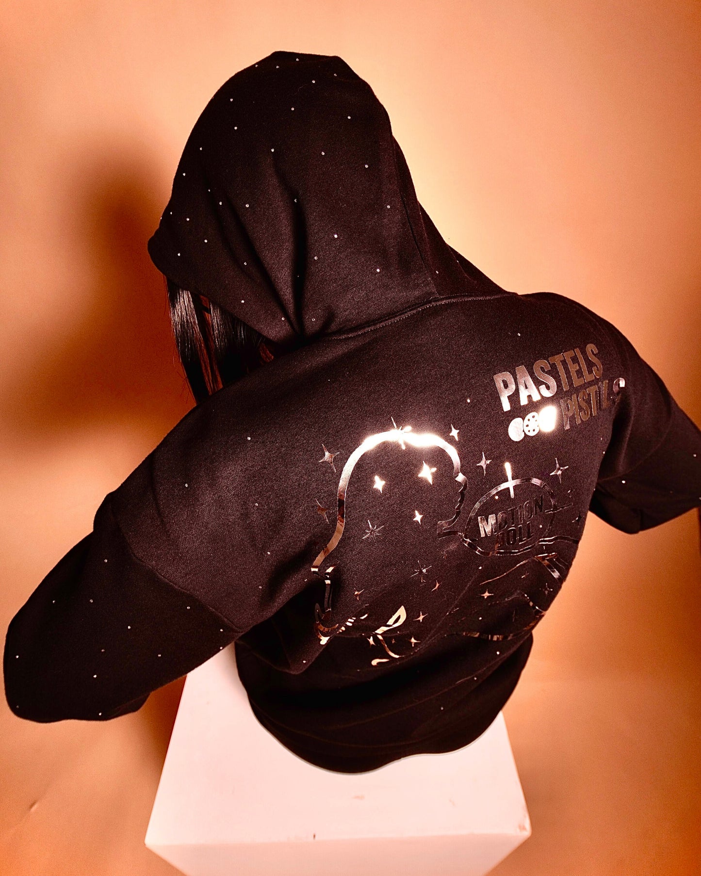 Back view of the Pastels & Pistols MotionDoLL Black Rhinestone Hoodie. Chrome vinyl back graphic and rhinestone layout visible. Warm studio lighting. Womens handmade hoodie.