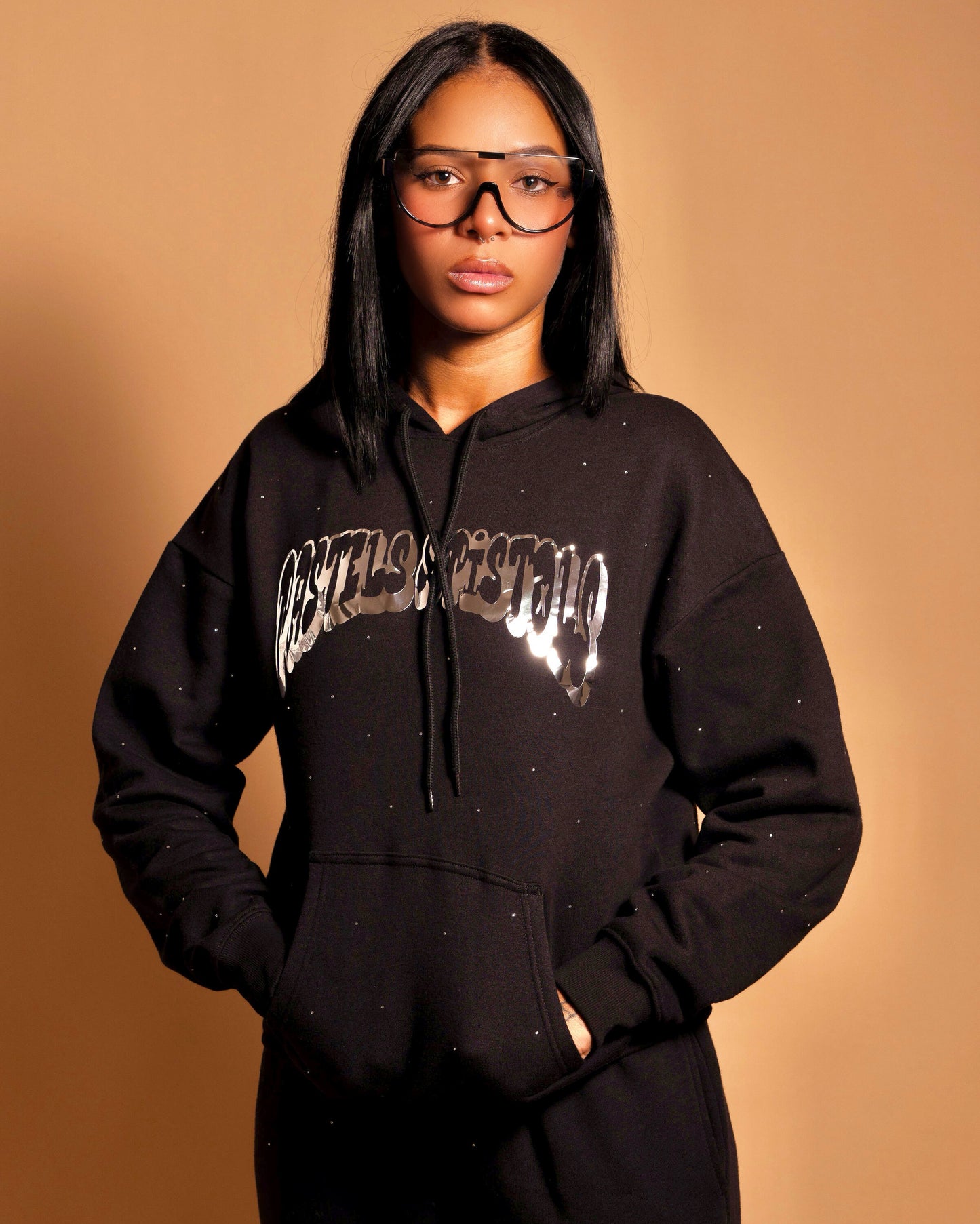 @yoselinpacheco394 wearing the Pastels & Pistols MotionDoLL Black Rhinestone Sweatsuit. Full front view of the handmade hoodie and jogger set with chrome vinyl logo and hand-placed rhinestones. Warm studio backdrop. Womens streetwear.
