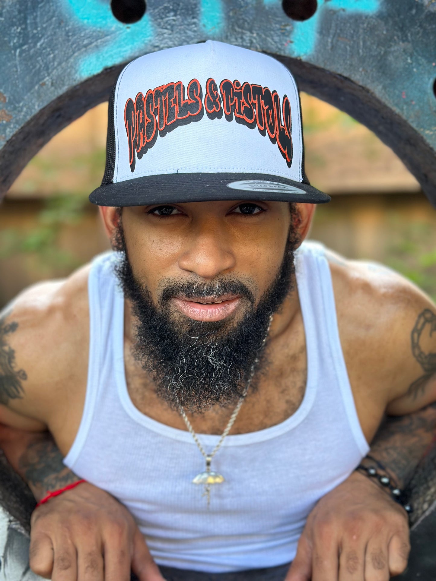 @jaethejewelerugz wearing the Pastels & Pistols Mother Puffer Flat Bill Trucker Hat with a white tank top. White front panel and red embroidered logo. Outdoor natural background with greenery.
