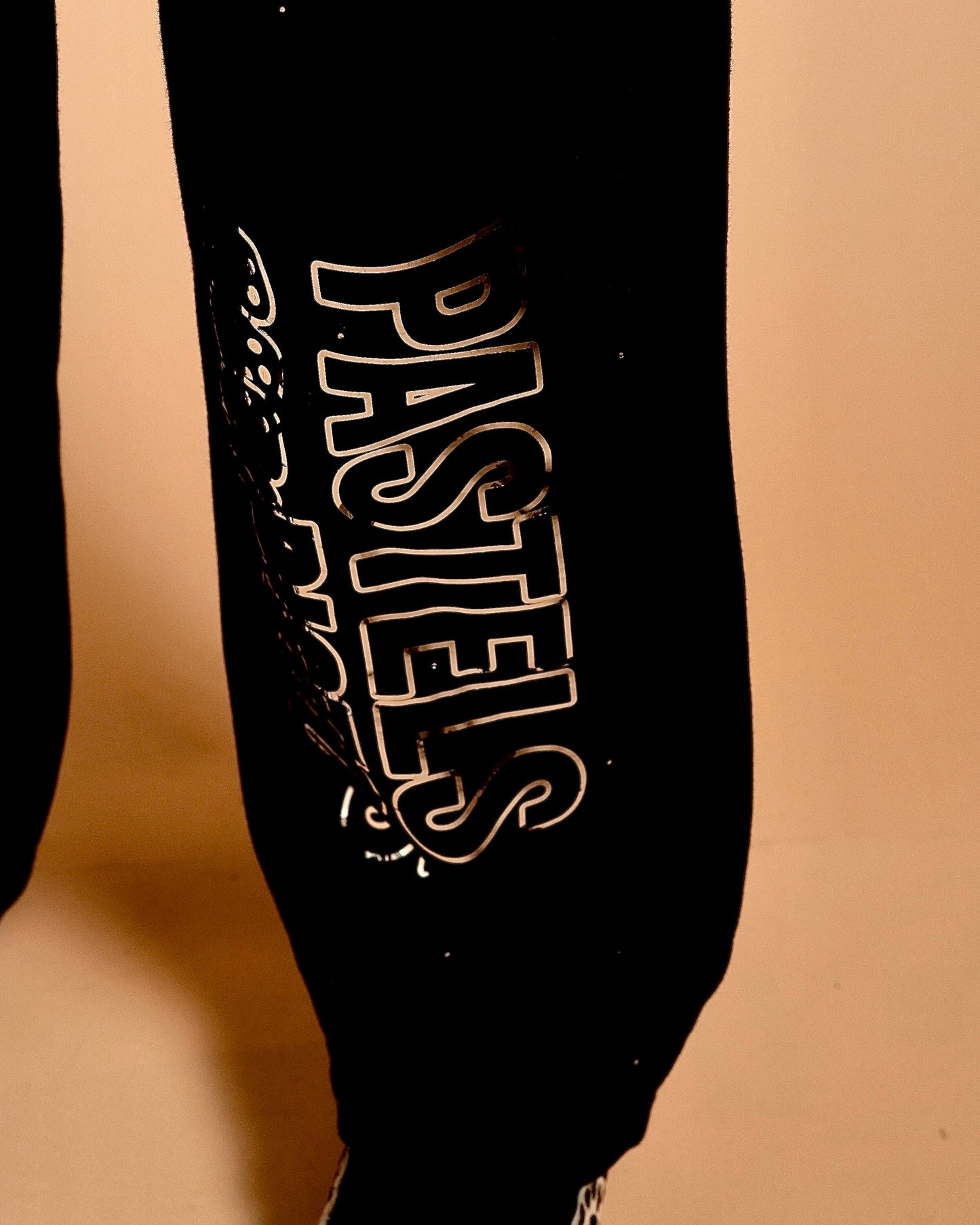 Side close-up of the Pastels & Pistols MotionDoLL Black Rhinestone Joggers. Chrome vinyl “PASTELS” leg print on black fabric. Warm studio backdrop. Womens jogger set.