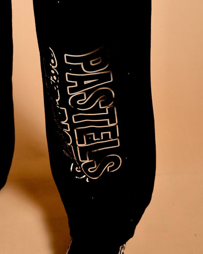 Side close-up of the Pastels & Pistols MotionDoLL Black Rhinestone Joggers. Chrome vinyl “PASTELS” leg print on black fabric. Warm studio backdrop. Womens jogger set.