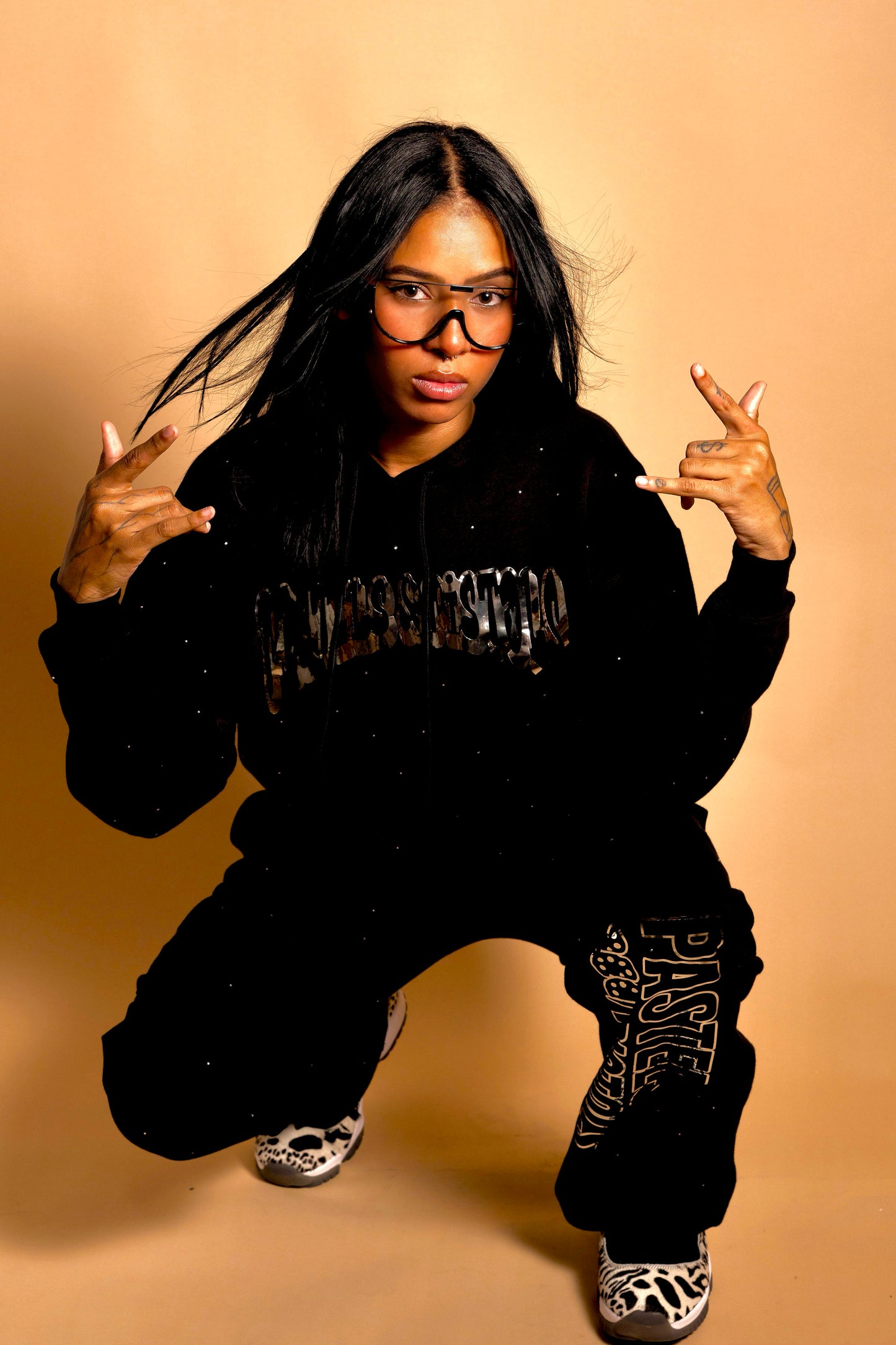 @yoselinpacheco394 in the Pastels & Pistols MotionDoLL Black Rhinestone Sweatsuit in an action pose. Full hoodie and jogger set with chrome vinyl graphics and rhinestones. Warm studio backdrop. Womens streetwear set.